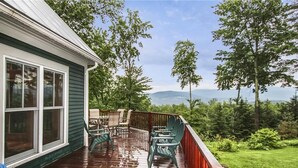 A wooden deck with outdoor furniture, a view of trees and mountains, and a house with green siding. - Treetops: 4 br, woodstove, a/c, deck & views, firepit, garage, great location! (Stowe)