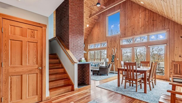 A spacious room with a wooden staircase, a fireplace, a dining area with wooden furniture, and large windows offering a view of the outdoors. - Treetops: 4 br, woodstove, a/c, deck & views, firepit, garage, great location! (Stowe)