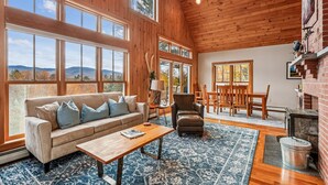 A spacious living room with a large sofa, a wooden coffee table, and a fireplace. - Treetops: 4 br, woodstove, a/c, deck & views, firepit, garage, great location! (Stowe)