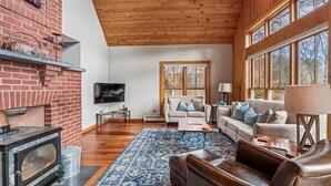 A cozy living room with a fireplace, a television, and a large window offering a view of trees. - Treetops: 4 br, woodstove, a/c, deck & views, firepit, garage, great location! (Stowe)