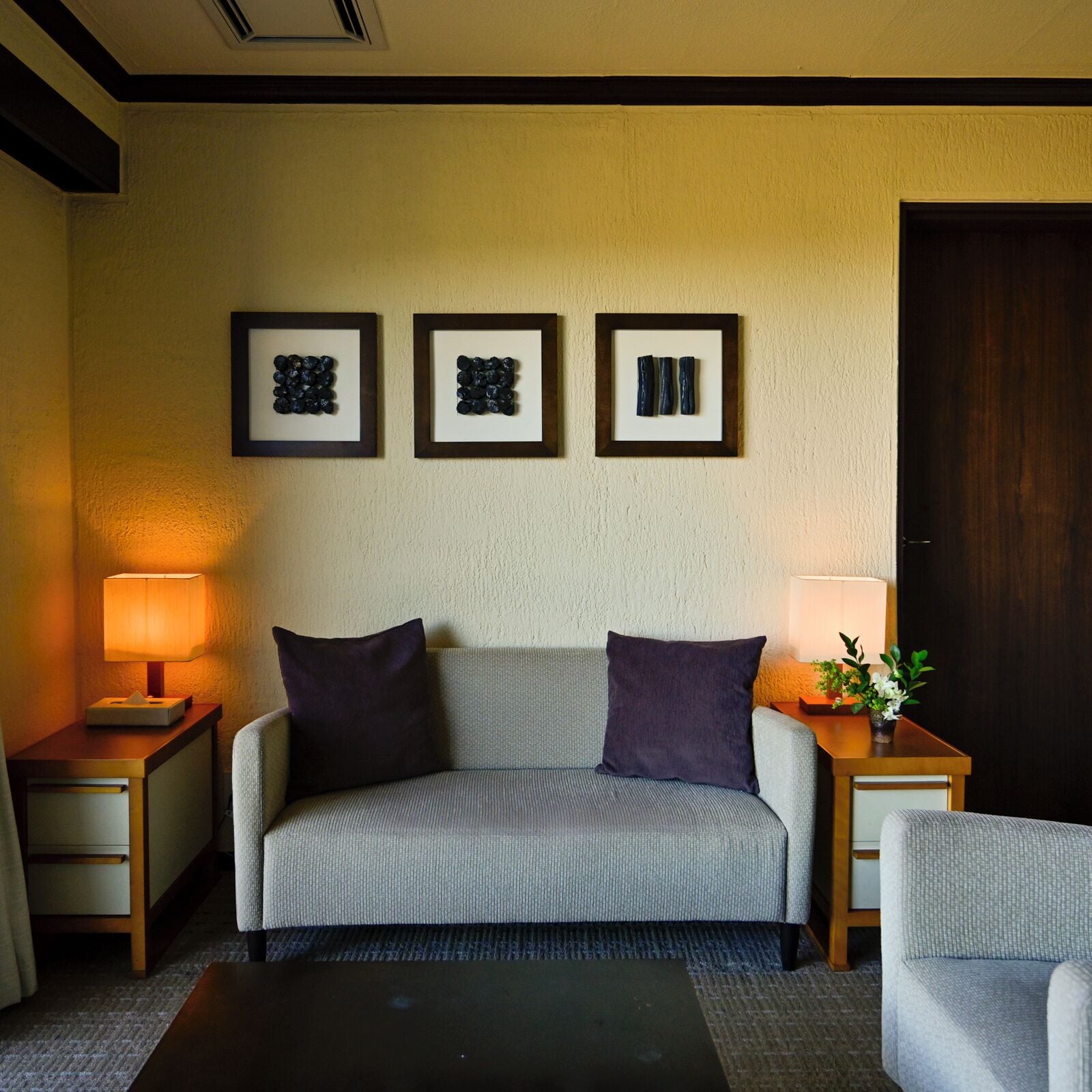 A hotel room with a sofa, two side tables with lamps, and three framed artworks on the wall.