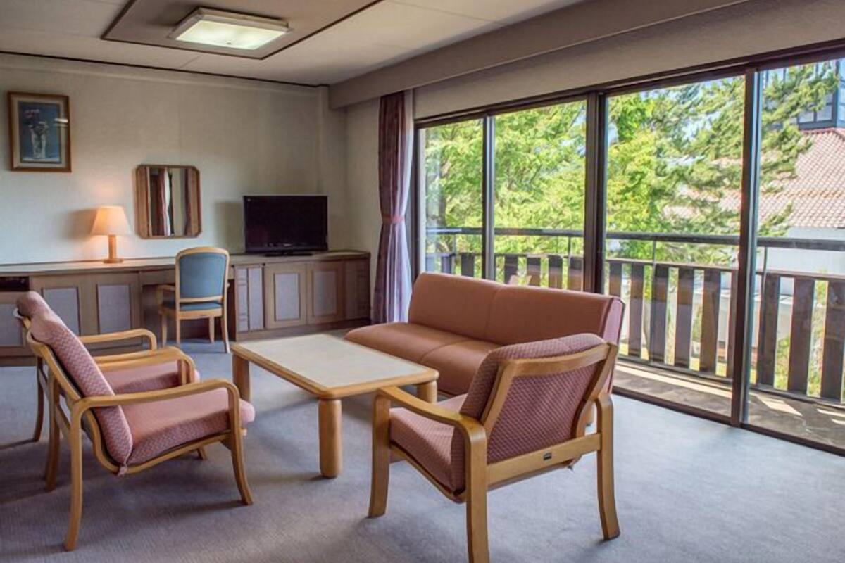 A room with a sofa, two armchairs, a small table, a TV, and a balcony with a railing.