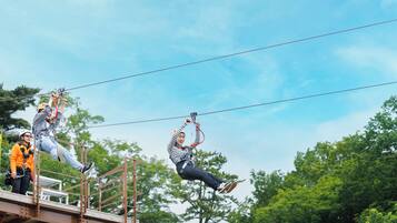 Una persona fa zip-line con l'attrezzatura di sicurezza, una piattaforma metallica e alberi sullo sfondo.