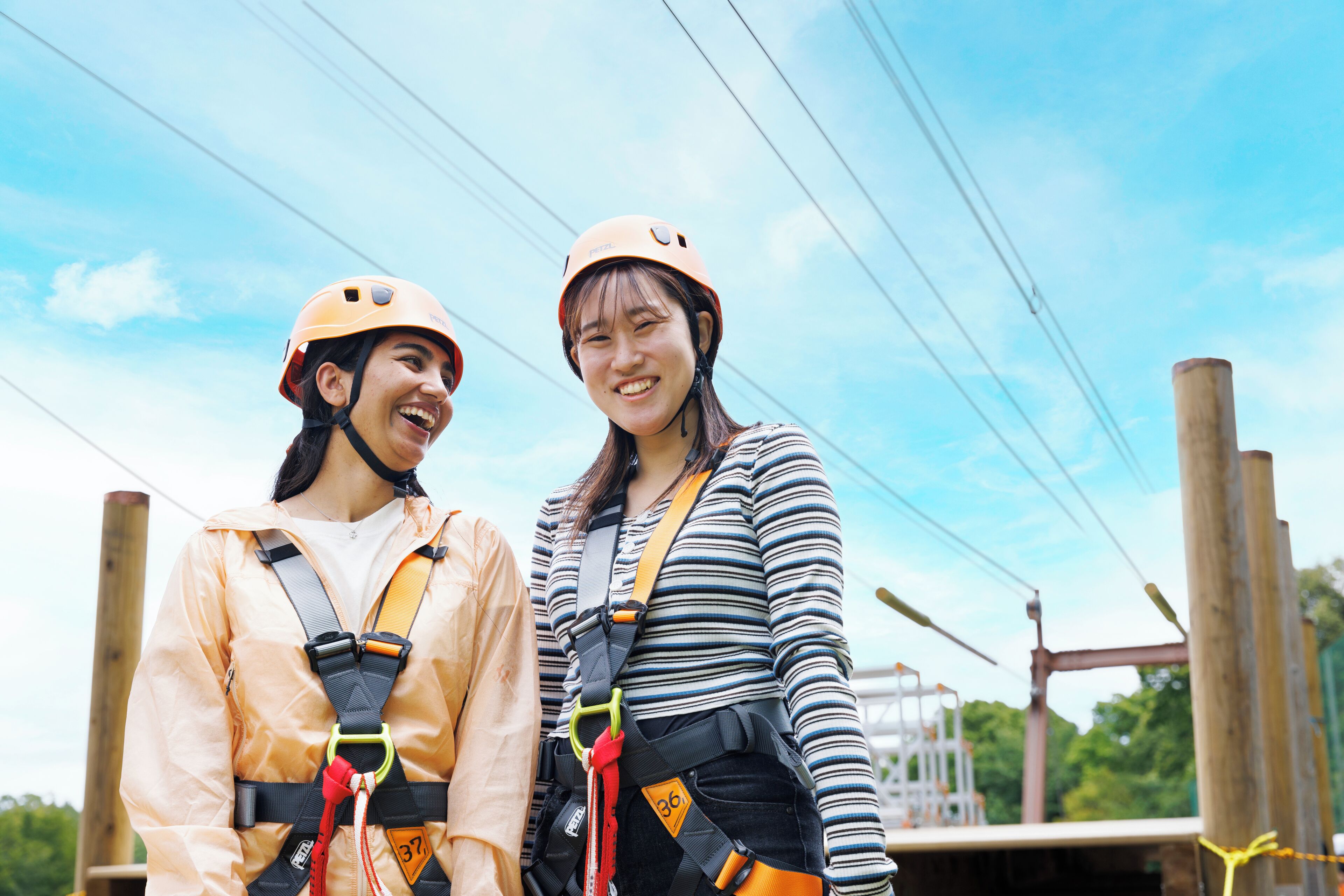 Two people wearing helmets and harnesses, standing on a wooden platform with a clear sky in the background.