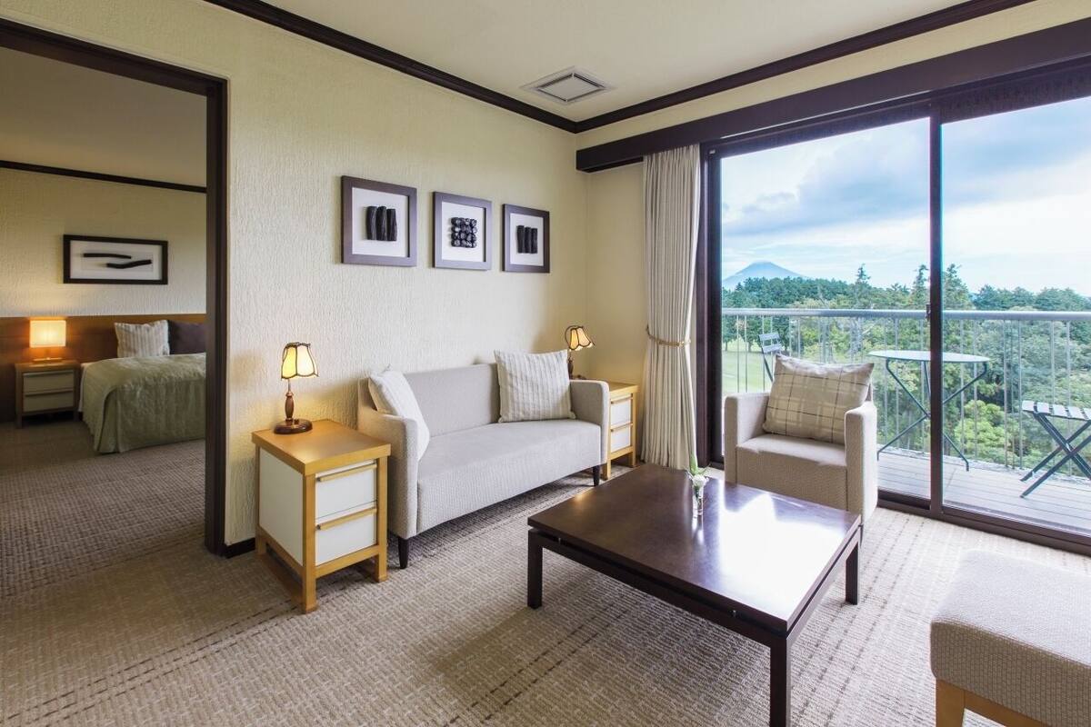 A hotel room with a sofa, a small table, a chair, a nightstand, and a bed. There are framed pictures on the wall and a view of the outdoors through large windows.