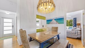 A dining area with a wooden table and wicker chairs, a hanging light fixture, and a painting of a pier on the wall. - AM I Dreaming: Heated Pool, Family Friendly Home, 3-min walk to beach! (Holmes Beach)