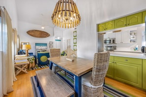 Uma cozinha compacta com armários verdes, uma mesa de jantar de madeira, cadeiras de vime e um lustre.