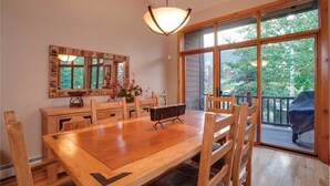 Un comedor con una mesa y sillas de madera, una araña, un espejo grande y vistas al exterior a través de amplios ventanales.
