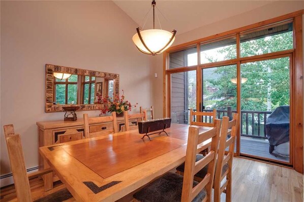 A dining area with a wooden table and chairs, a chandelier, a large mirror, and a view of the outdoors through large windows.