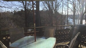 Une terrasse extérieure avec une table en verre et des chaises en métal, donnant sur une étendue d’eau et des arbres.