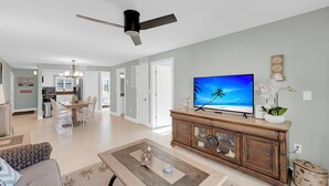 Una sala de estar con televisor, ventilador de techo, comedor y cocina al fondo.