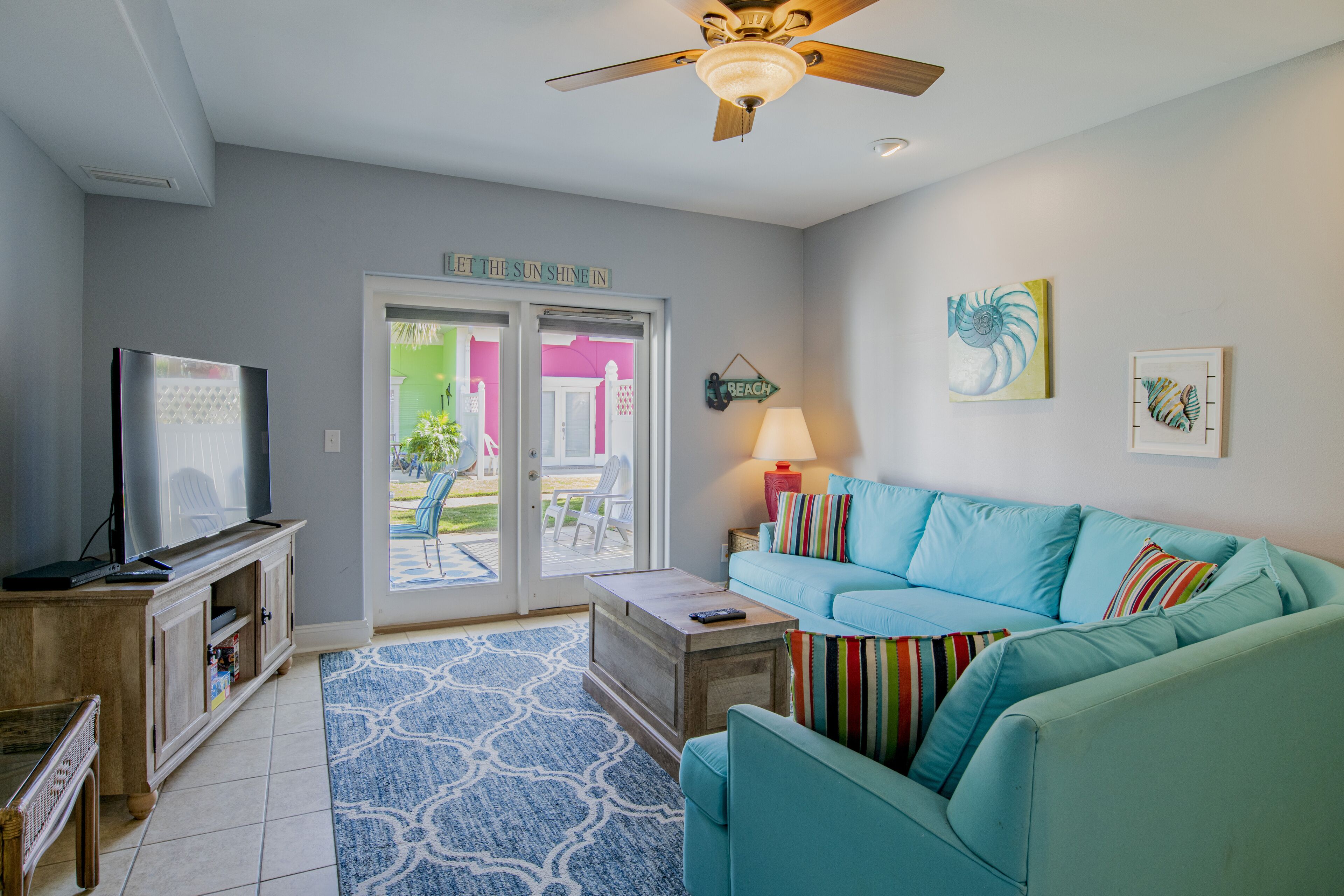 A living room with a teal sofa, a television on a wooden cabinet, a ceiling fan, and a door leading outside.