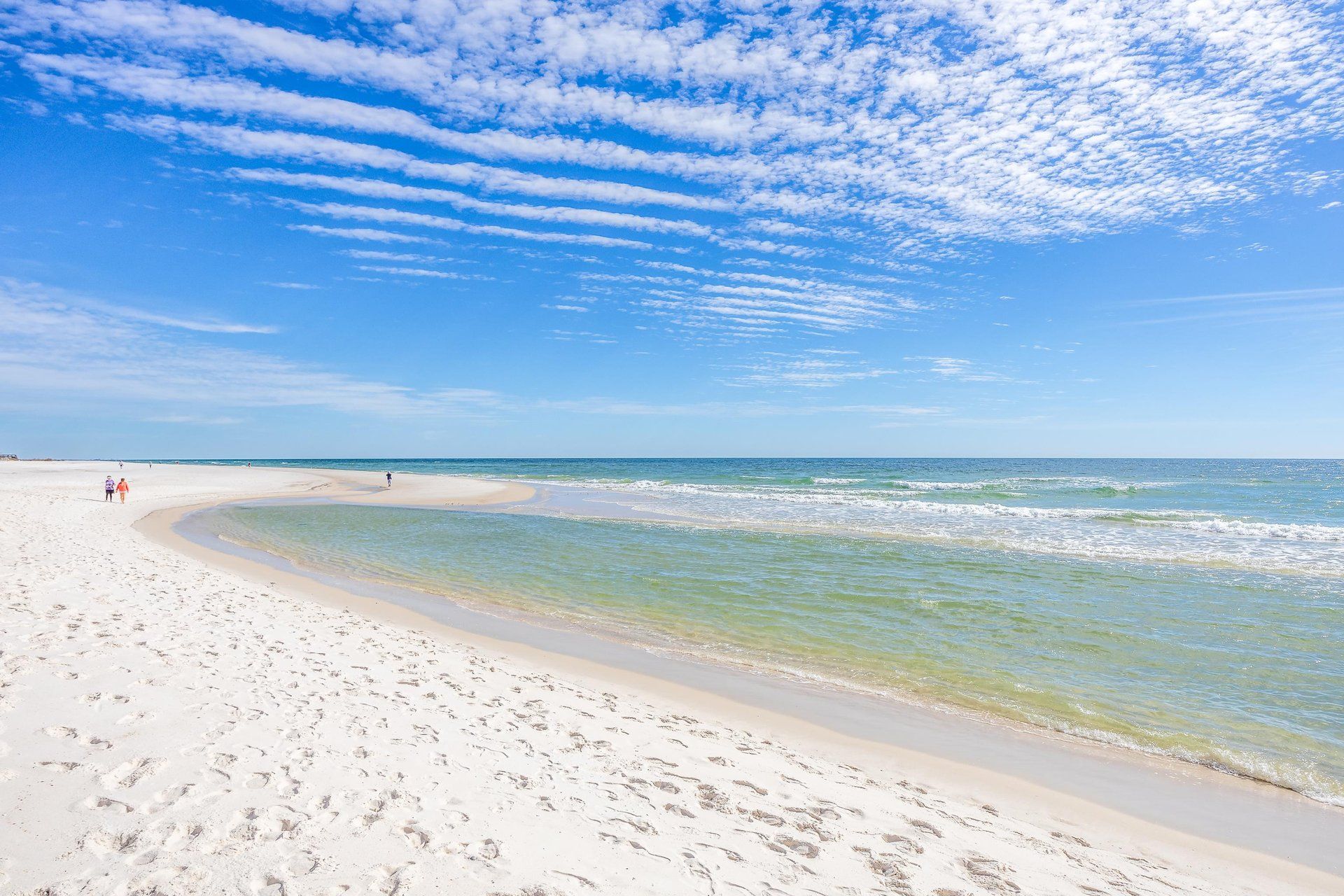 A wide sandy beach with clear blue waters and a few people walking.
