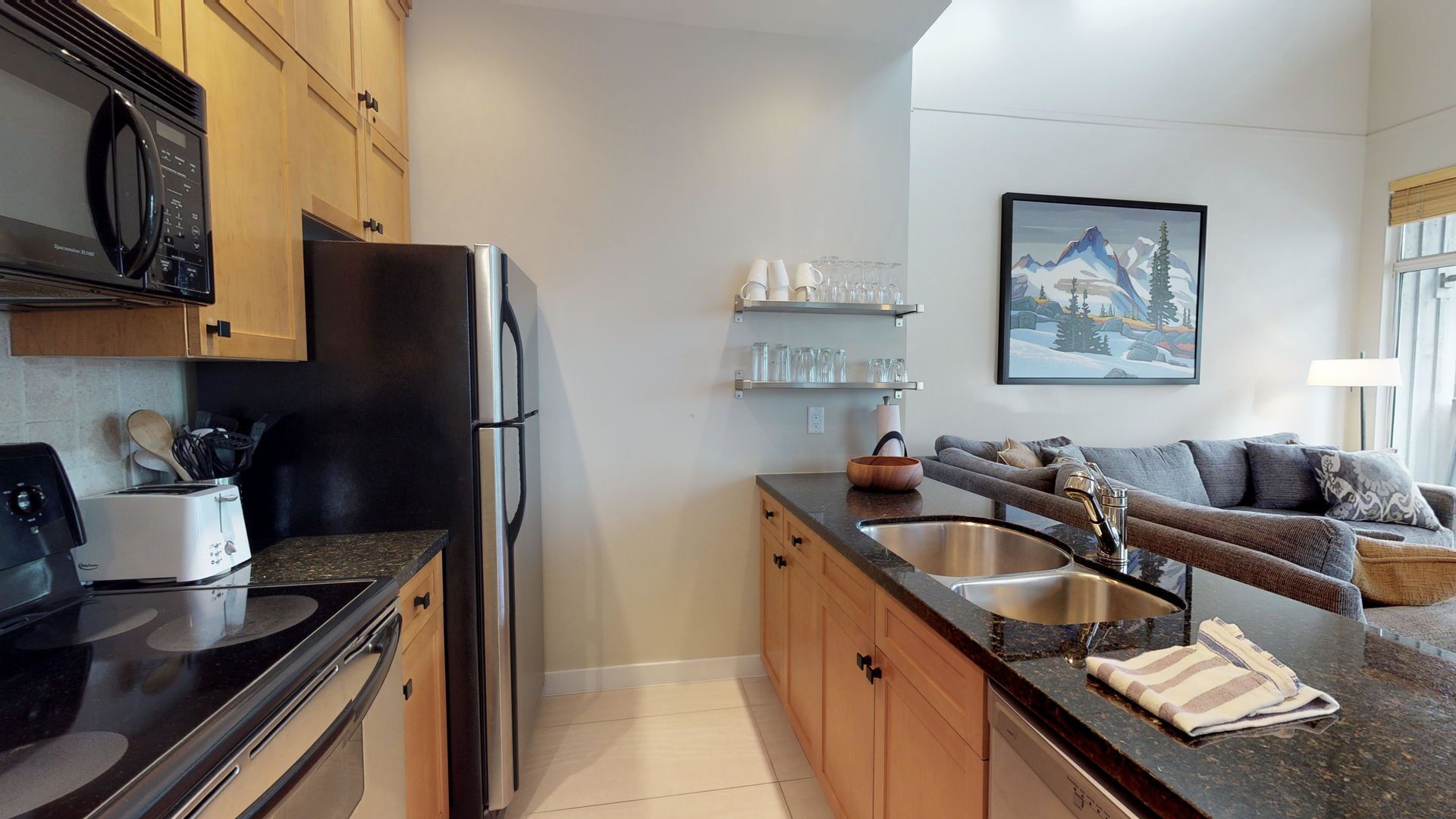 A modern kitchen with a refrigerator, stove, and sink. There is a dining area with a sofa and a painting on the wall.
