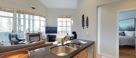 A modern kitchen with a granite countertop, stainless steel sink, and a dishwasher. There is a dining area with a table and chairs, and a living room with a sofa and a TV.