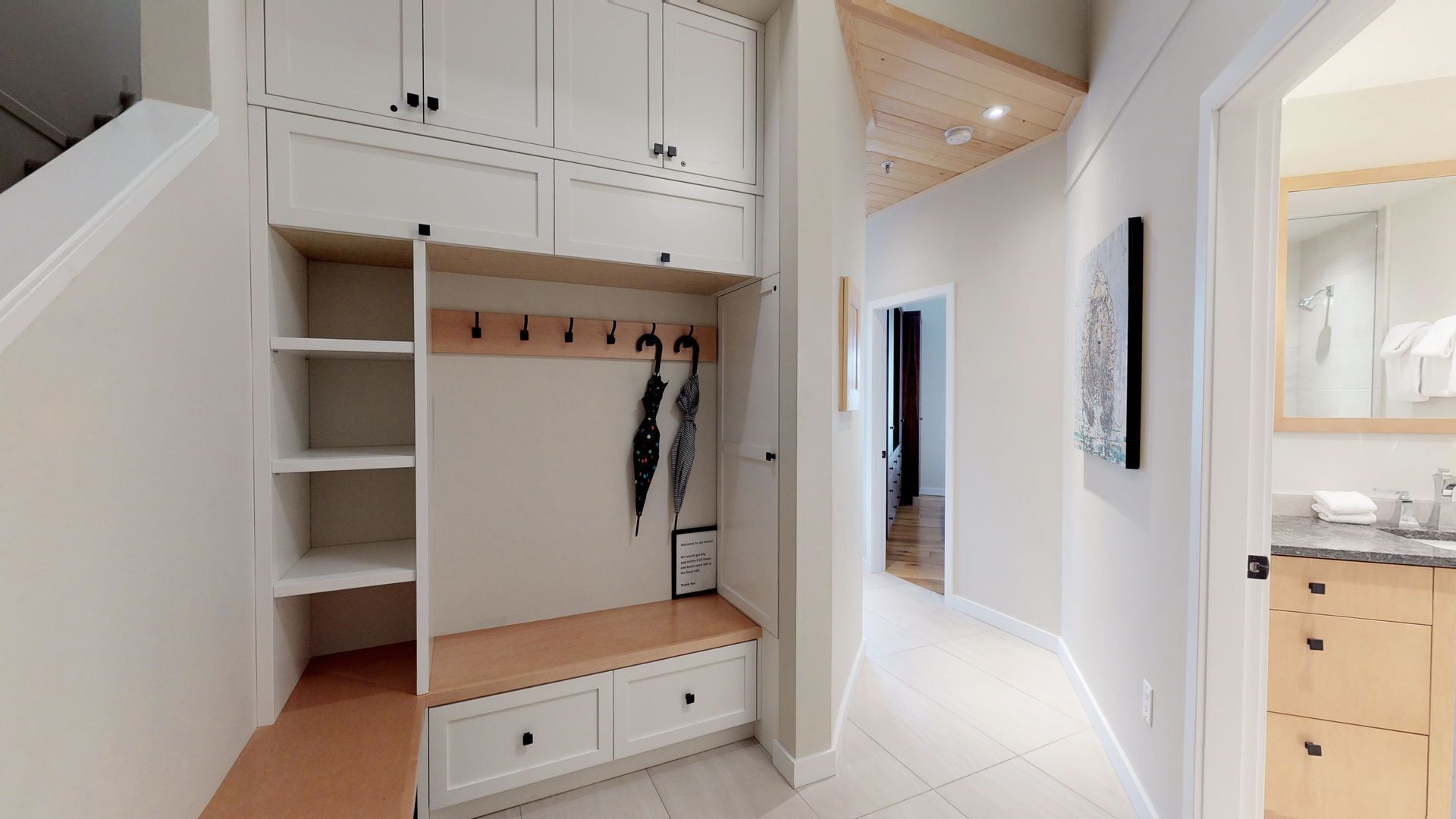A modern interior with a built-in wardrobe, a wooden bench with hooks, and a bathroom with a mirror and sink.