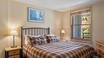 Um quarto com uma cama, uma mesa de cabeceira com luminária, cômoda, janela com vista para árvores e um quadro na parede.