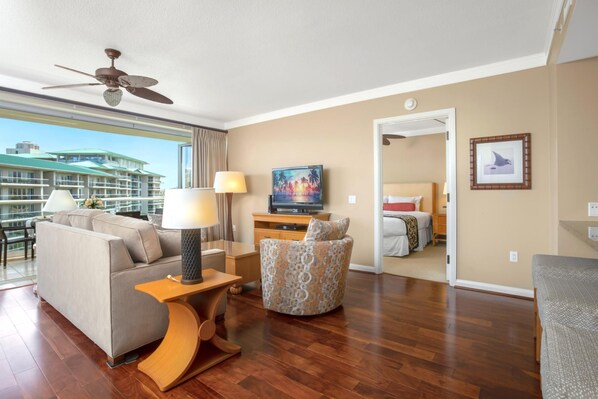 A hotel room with a balcony, a sofa, a patterned armchair, a TV, and a bed.