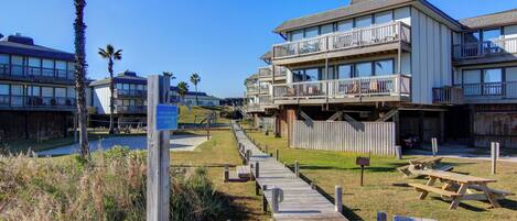 A wooden boardwalk leading to a beachfront hotel with multiple balconies and picnic tables.