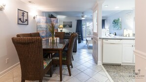 A dining area with wicker chairs and a glass table, a kitchen with white cabinets, and a view to the outside.