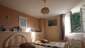 Une chambre avec deux lits simples, une étagère avec des livres et des photos, une fenêtre avec des rideaux et un luminaire au plafond.