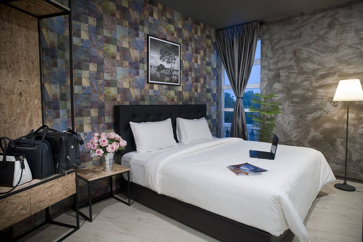 A neatly arranged hotel room with a bed, a bedside table with a lamp, a desk with bags, and a wall with a patterned design.