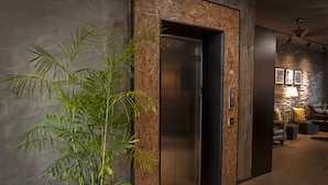 Un ascenseur moderne avec un revêtement mural en bois, un pot de bambou placé à côté, et un coin salon avec une lampe et un canapé en arrière-plan.