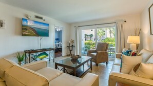 Uma sala de estar com sofá, poltrona, mesa de centro e um quadro de uma paisagem costeira.