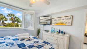 Quarto com uma cama grande, janela com persianas, televisão, painel decorativo na parede e cômoda branca com vasos azuis.