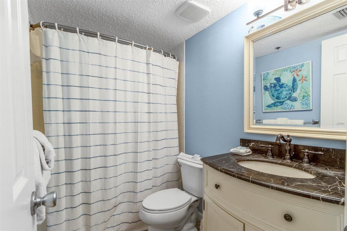 Een badkamer met een gestreept douchegordijn, een wit toilet, een wastafel met een marmeren blad en een spiegel met een schilderij van een zeeschildpad.