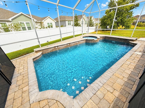 A rectangular swimming pool with a stone border and a hot tub in the center, surrounded by a white fence and string lights.