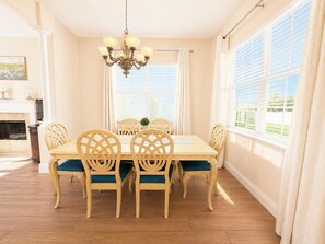 A dining room with a wooden table and chairs, a chandelier, and large windows with blinds.