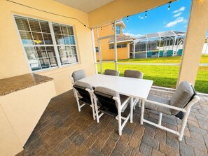 A covered patio area with a table and chairs, a window with blinds, and a view of a house and lawn.