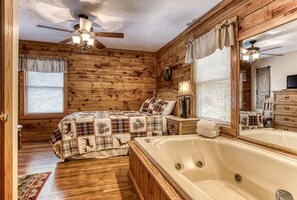 A log cabin bedroom with a large bed, a wooden headboard, a nightstand with a lamp, a large mirror, and a bathtub. - ER13 - Moose Manor  Great location - Close to town! (Pigeon Forge)