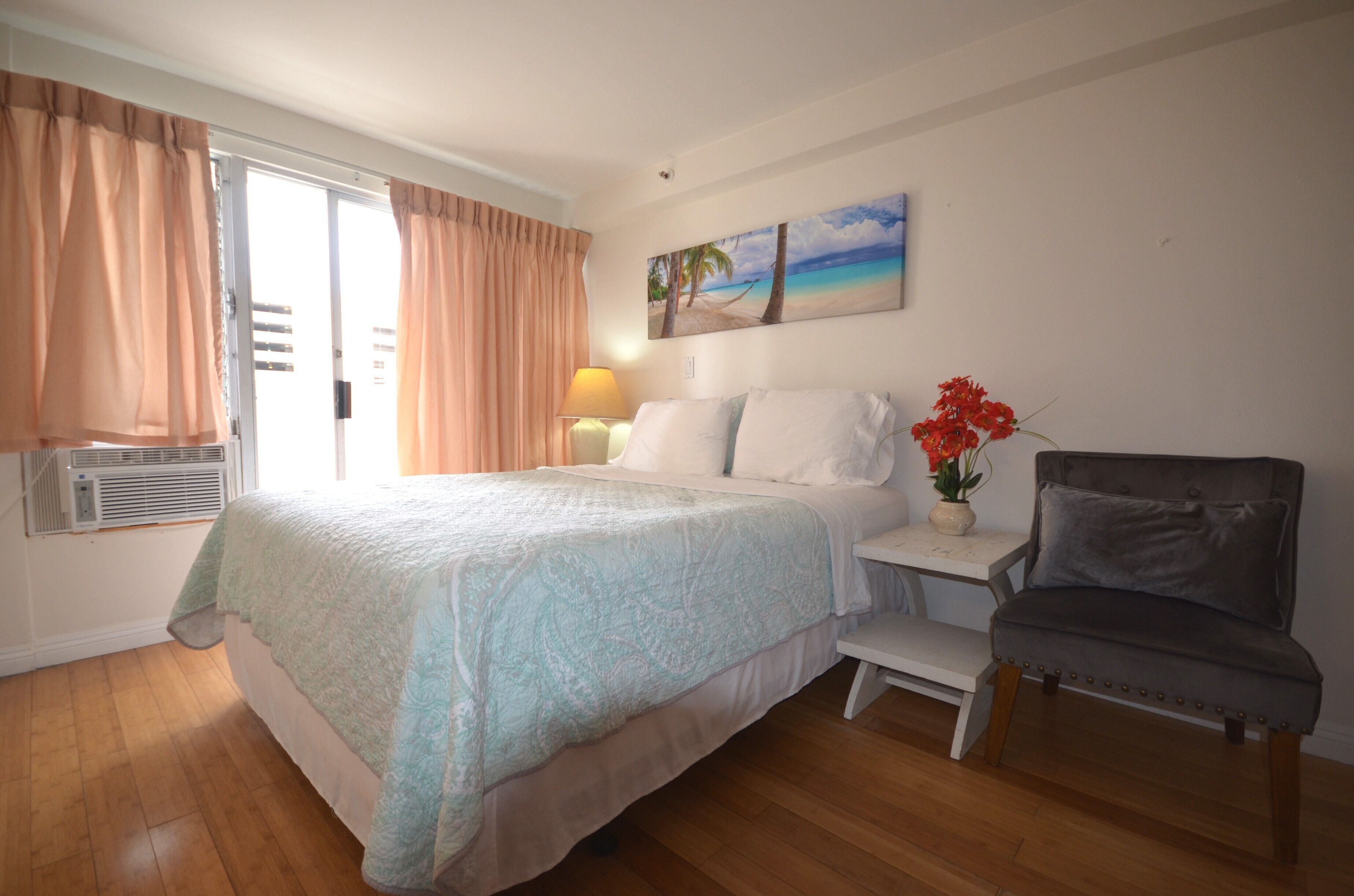 Een slaapkamer met een bed, een stoel, een nachtkastje, een lamp en een schilderij van een strand.