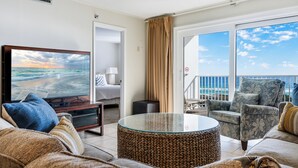 A hotel room with a TV displaying a beach scene, a balcony with a view of the ocean, and a seating area with a wicker ottoman. - 🌊 Islander #506 – Affordable Gulf-View Getaway! 🌴✨ (Destin)