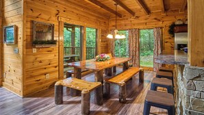 A wooden cabin interior with a large dining table, benches, a stone fireplace, and a view of the forest through large windows. - Family Fun | Theater, Gameroom, Hot Tub (Gatlinburg)