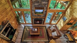A cozy cabin interior with a fireplace, wooden furniture, and large windows offering a view of the surrounding forest. - Family Fun | Theater, Gameroom, Hot Tub (Gatlinburg)