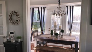 A dining area with a wooden table, chairs, a chandelier, and a view of the outdoors through large windows with black and white curtains.