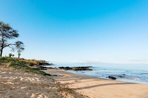 Una spiaggia sabbiosa con un cielo azzurro limpido, affioramenti rocciosi e vegetazione rada.