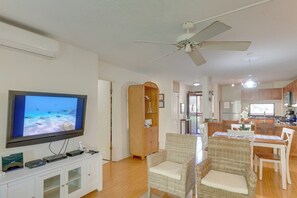 A living room with a flat-screen TV, a ceiling fan, wicker furniture, and a kitchen in the background.