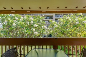 Un balcone con un tavolino in vetro e due sedie in vimini, con vista su un edificio con balconi e alberi fioriti di bianco.