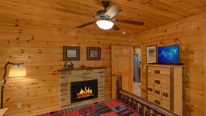 A log cabin interior with a fireplace, a bed with a red and black quilt, a ceiling fan, and a television on a wooden cabinet. - Natural Attractions: Theater, Gameroom, Hot Tub (Gatlinburg)