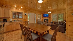 A cabin interior with a wooden dining table, chairs, a sofa, a fireplace, a television, and a kitchen area. - Natural Attractions: Theater, Gameroom, Hot Tub (Gatlinburg)