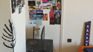 Ett rum med en vägg täckt av olika affischer och bilder, ett skrivbord med en dator och en lampa samt en säng med en filt.