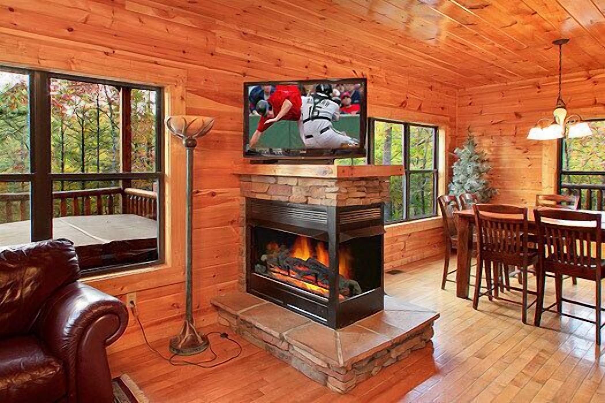 A wooden cabin interior with a stone fireplace, a television mounted above it, and a dining area with wooden furniture.