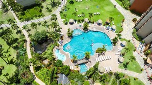 An aerial view of a resort with a central swimming pool, surrounded by palm trees, lounge chairs, and a small waterfall.