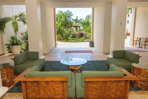 Um saguão com sofás verdes, uma mesa de centro e vista para um jardim tropical.
