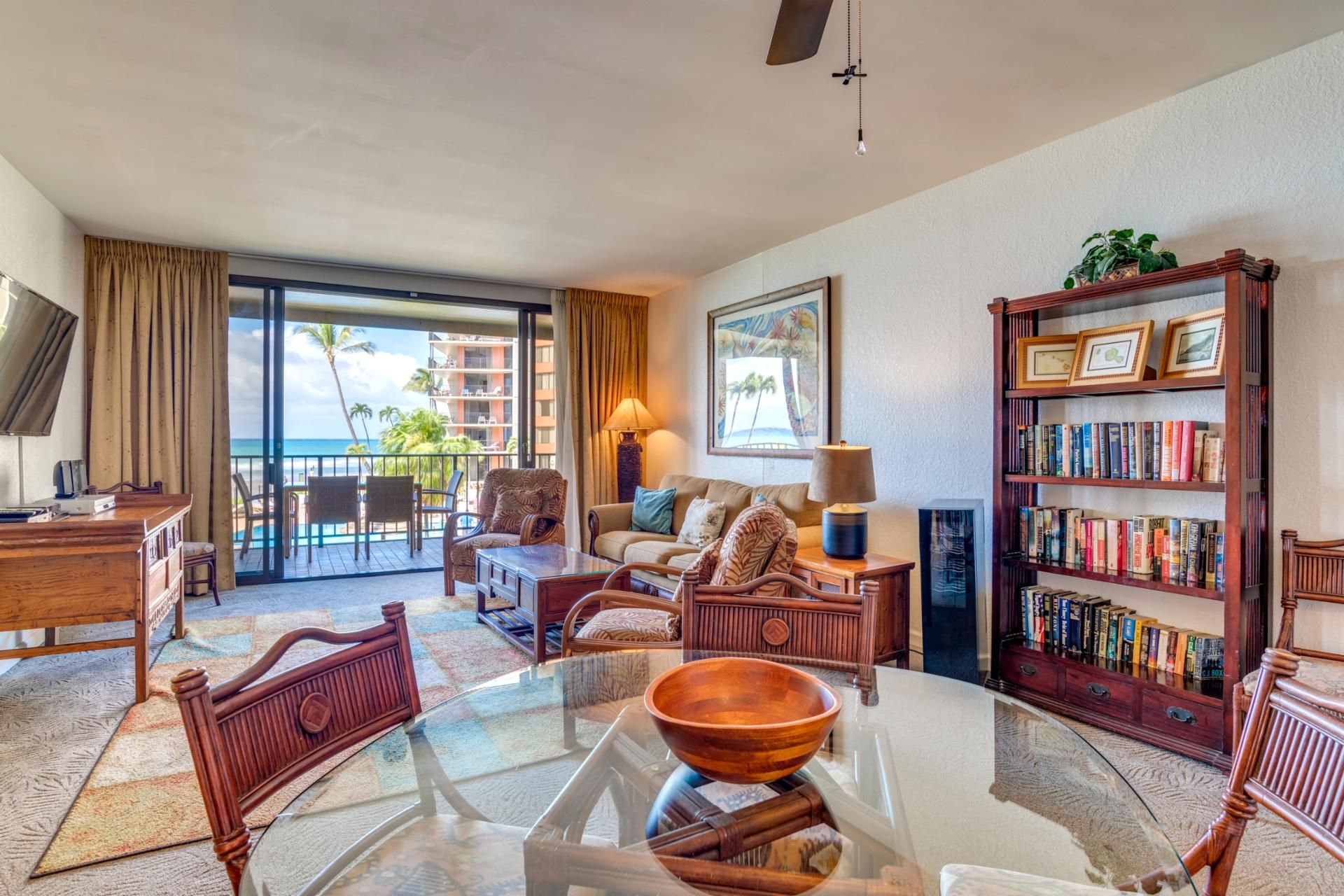A spacious living room with a glass coffee table, comfortable seating, a bookshelf, and a view of the ocean.