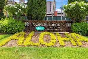 Placa do Restaurante & Bar Kaanapali Shores Beach Club com logotipo, cercada por vegetação e um muro de pedra.
